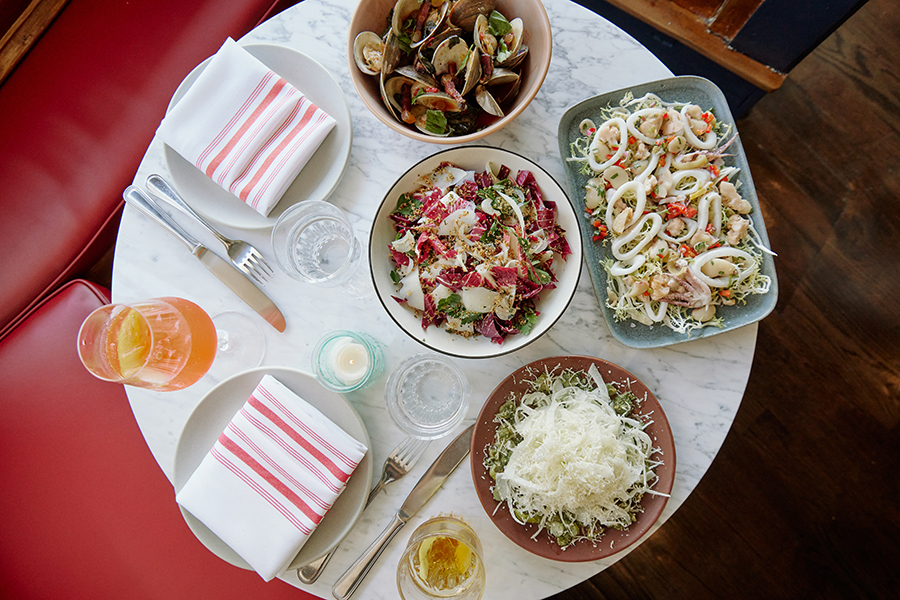 Overhead view of a circular white marble table covered with Italian-inspired salads, clams, and other small plates.