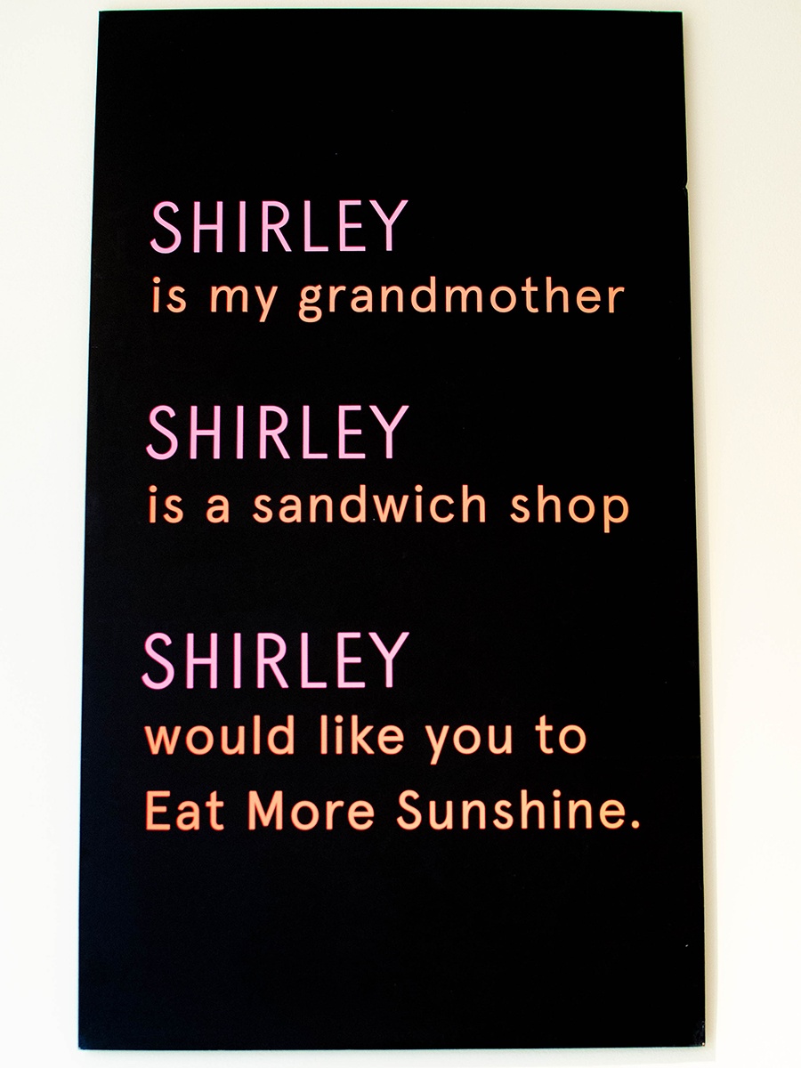 A black sign has orange and pink text reading: Shirley is my grandmother. Shirley is a sandwich shop. Shirley would like you to Eat More Sunshine.