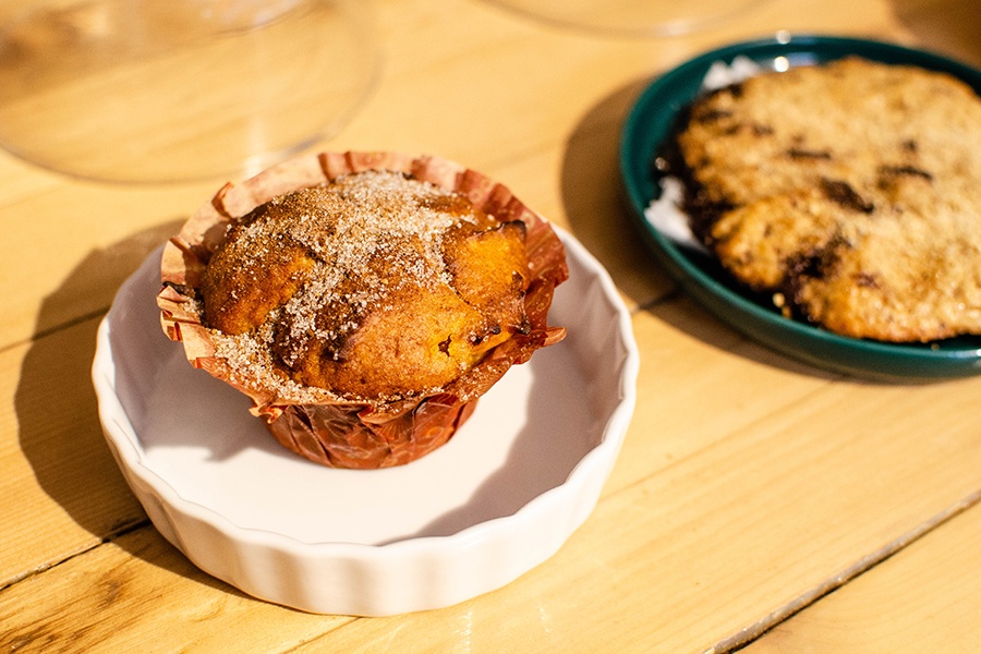 A golden brown muffin with a sugary topping sits in a small decorative plate with a cookie visible in the background.