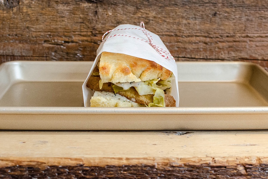 A tofu sandwich is wrapped in white paper and tied with a red and white string. It sits on a metal tray on a wooden counter.