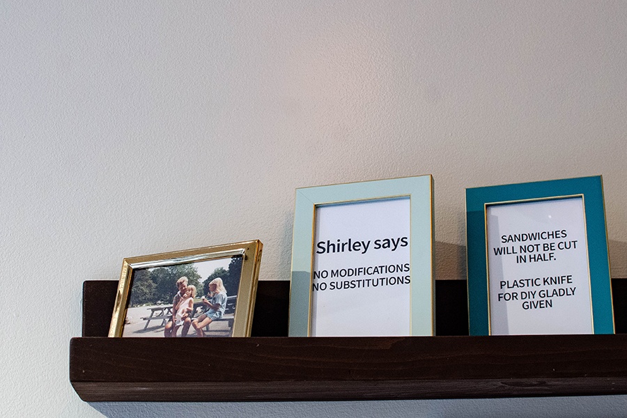 A shelf holds a photograph and two framed signs indicating that a restaurant offers no substitutions or modifications and doesn't cut sandwiches in half.