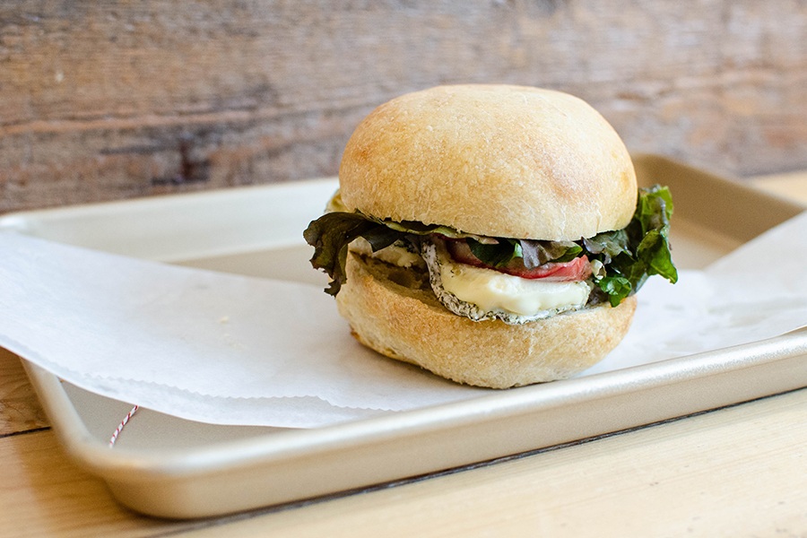 A sandwich on a round bun sits on a white paper on a silver tray. The sandwich is stuffed with a big hunk of cheese, greens, and slivers of red apple.
