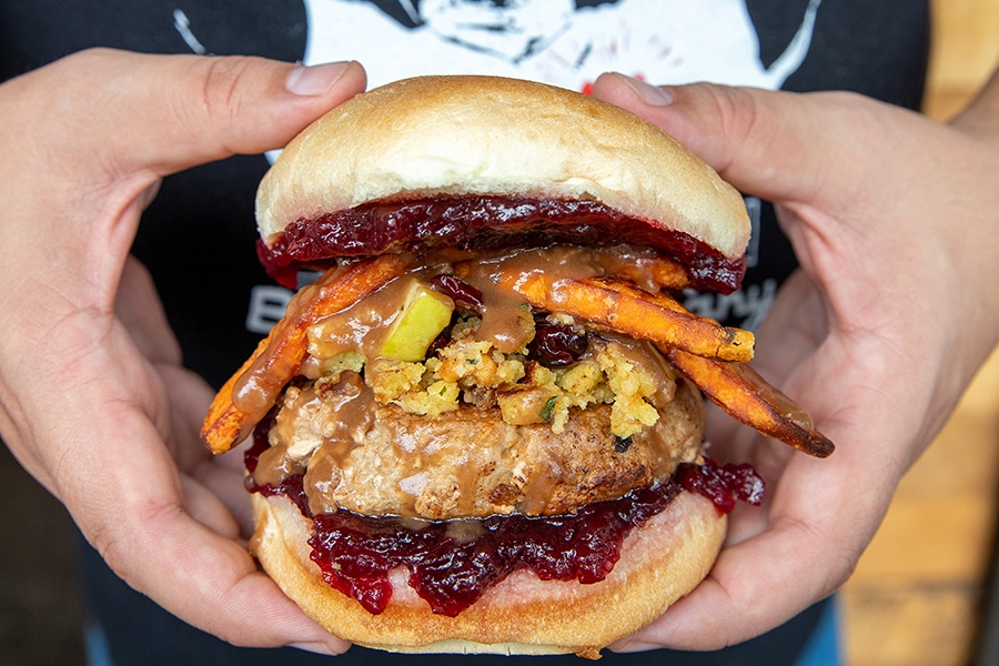 Closeup on a turkey burger held by two hands. It's topped with ample amounts of stuffing, cranberry sauce, and thin sweet potato fries.