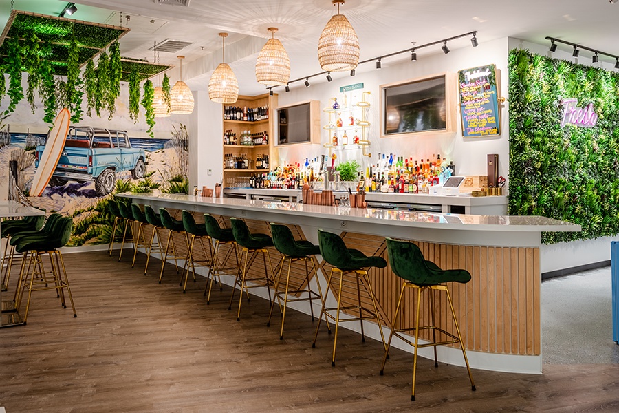 Interior shot of an empty, brightly lit bar with a large beach mural featuring a surfboard and a Jeep, plus light wood accents, shelves of liquor bottles, and a green plant wall with neon signage reading Fresh.