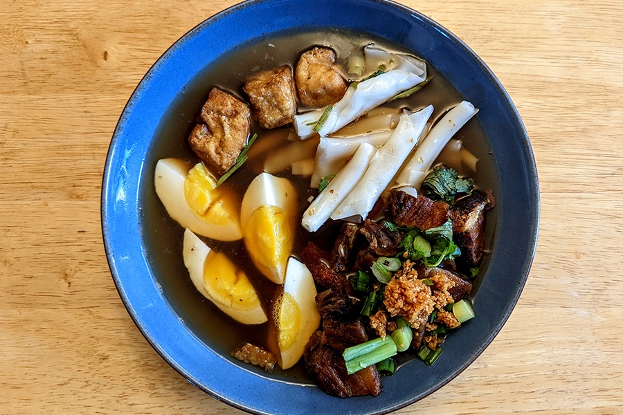 Overhead view of a bowl of soup with a dark broth, a boiled egg, pieces of fried tofu, crispy pork belly, and a scallion garnish.