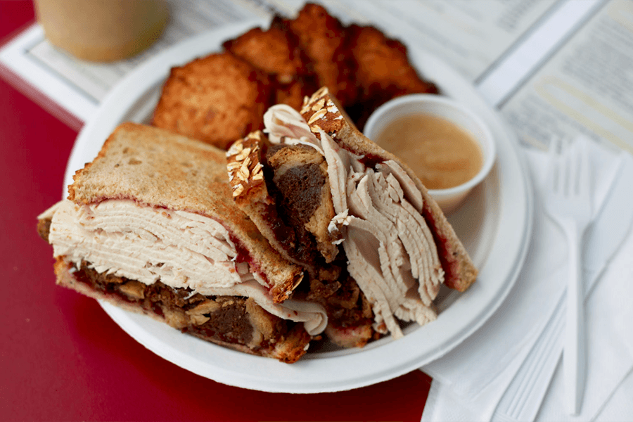 A deli sandwich stacked thick with turkey is sliced in half and served on a plate with potato latkes and a side of apple sauce.