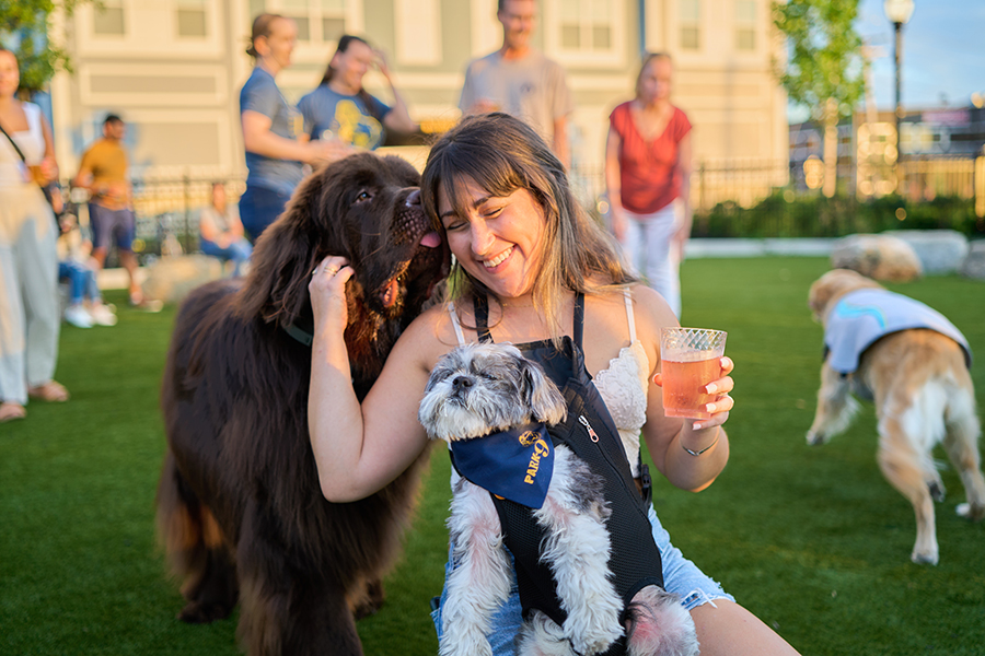 A woman holding a beer is wearing a small dog in a forward-facing carrier and smiling while petting another dog that is licking her face.