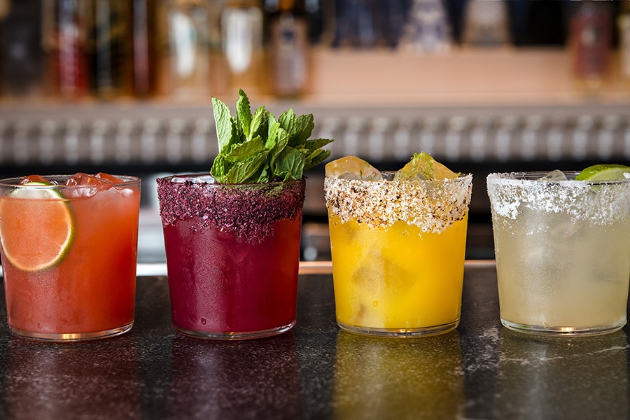 Four colorful cocktails with salt rims are lined up on a bar.