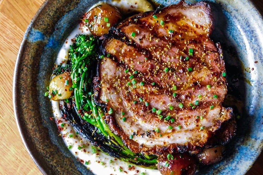 A grilled round of porchetta is garnished with chopped herbs and sits on a bed of charred broccolini and a white puree. 