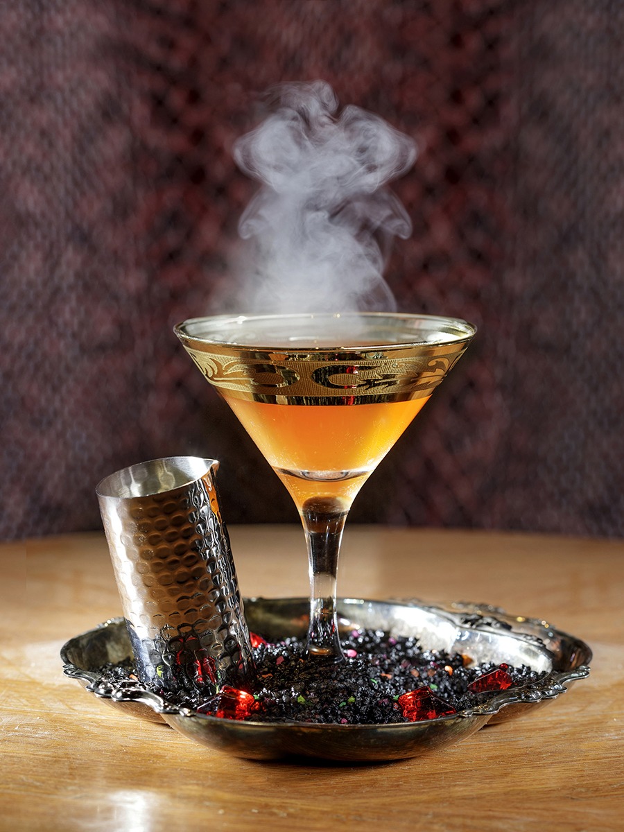 An orange-brown cocktail sits in a martini glass with smoke coming off the top.