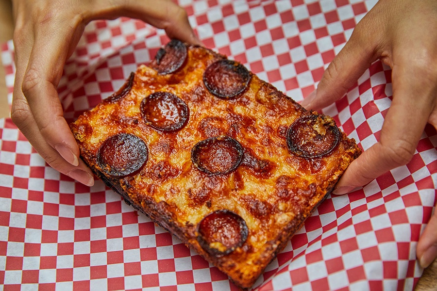 Hands hold a square slice of pepperoni pizza over red-and-white-checkered paper.