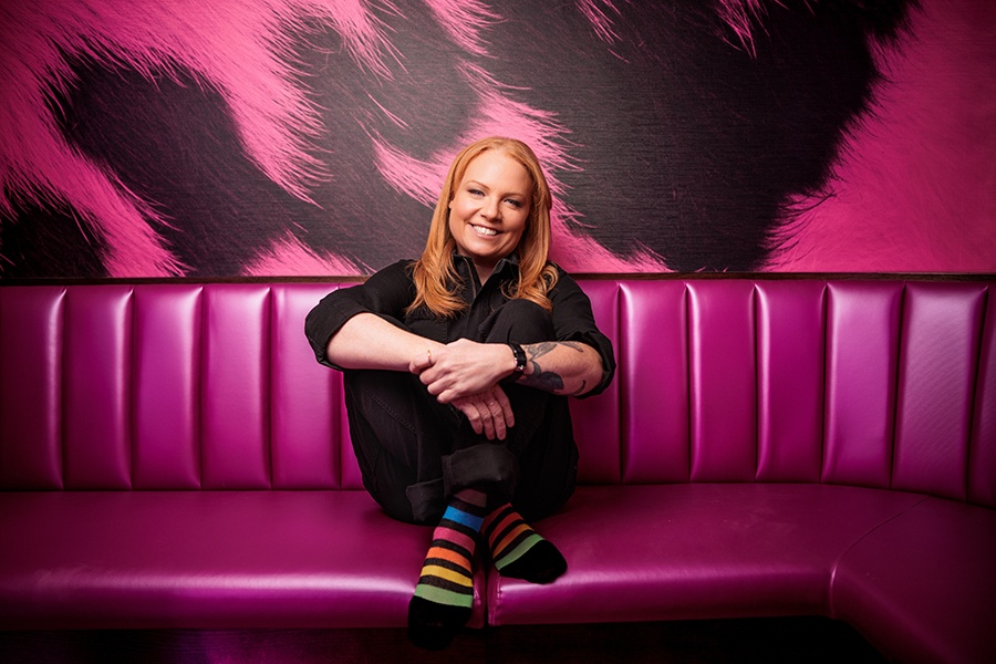 A red-haired, smiling woman in black clothing (except for rainbow-striped socks) sits on a bright pink couch in front of black and pink leopard print wallpaper.