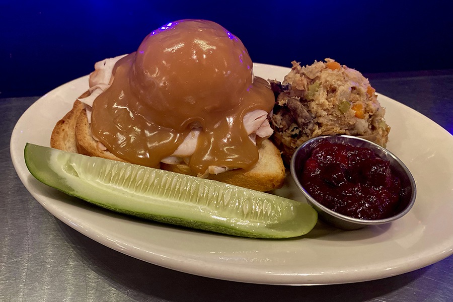 An open-faced sandwich features a big scoop of gravy-drenched mashed potatoes atop a slice of grilled bread, with a pickle, stuffing, and cranberry sauce on the side.