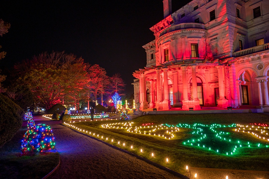 The exterior of an elegant mansion and the lawn in front of it are illuminated in strings of different colored Christmas lights.