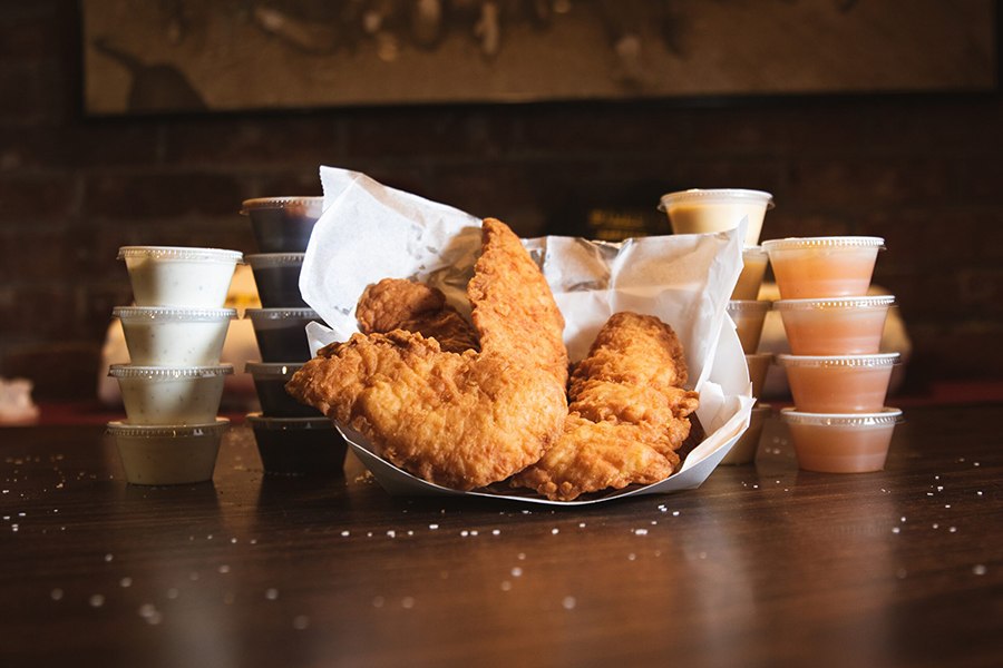 A paper basket full of fried chicken tenders is flanked by small plastic cups of four different dipping sauces.