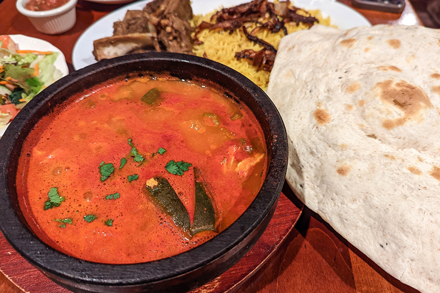 A bright red stew in a black bowl is accompanied by a large, round, naan-like bread. A roasted lamb and rice dish is visible in the background.