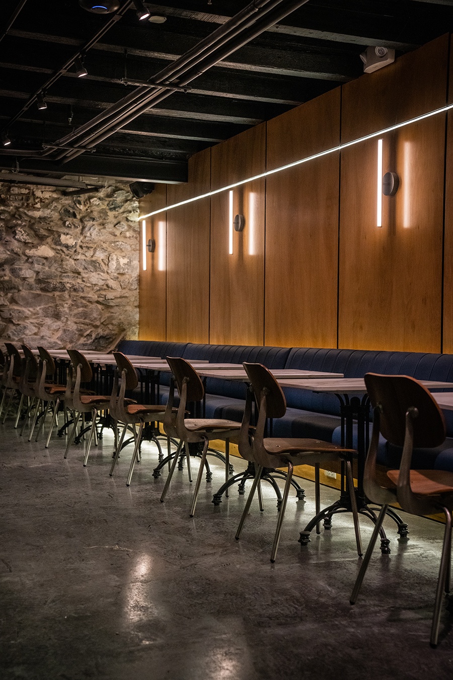 A blue banquette, plus tables and chairs, line an empty bar space with a stone wall and a wood-paneled wall.