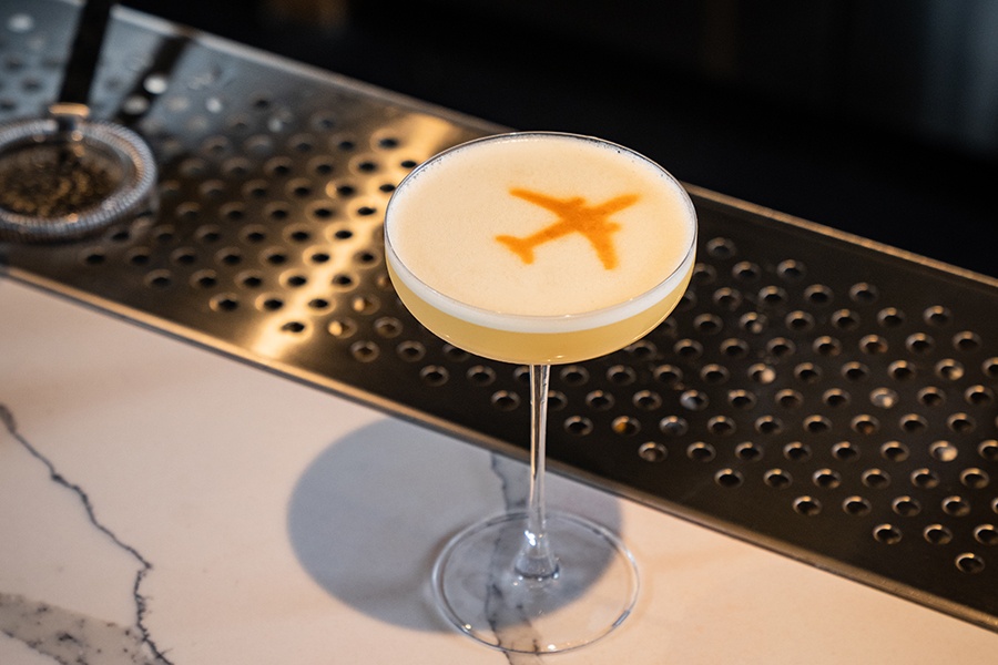 A pinkish cocktail in a martini glass has an airplane-shaped silhouette on top of the drink.