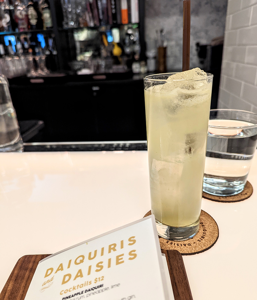 A pale yellow cocktail in a tall glass sits on a white bar next to a menu that says Daiquiris and Daisies.