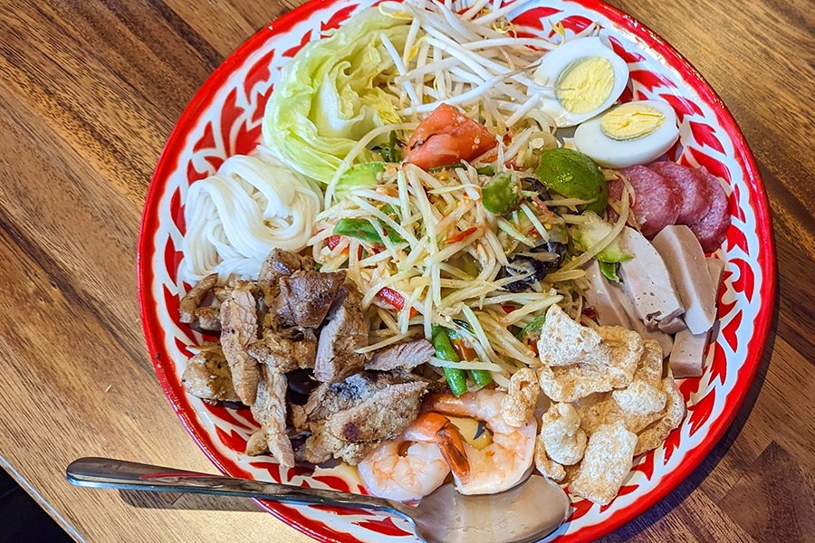 Overhead view of a Thai papaya salad garnished with shrimp, pork rinds, a hard-boiled egg, noodles, and more.