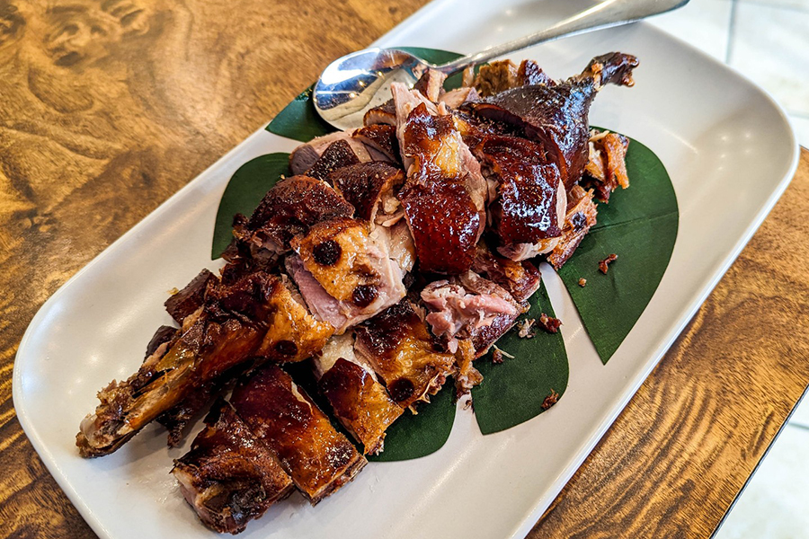 Sliced duck with a glossy reddish-brown crispy skin is spread across a decorative tropical leaf on a white plate.