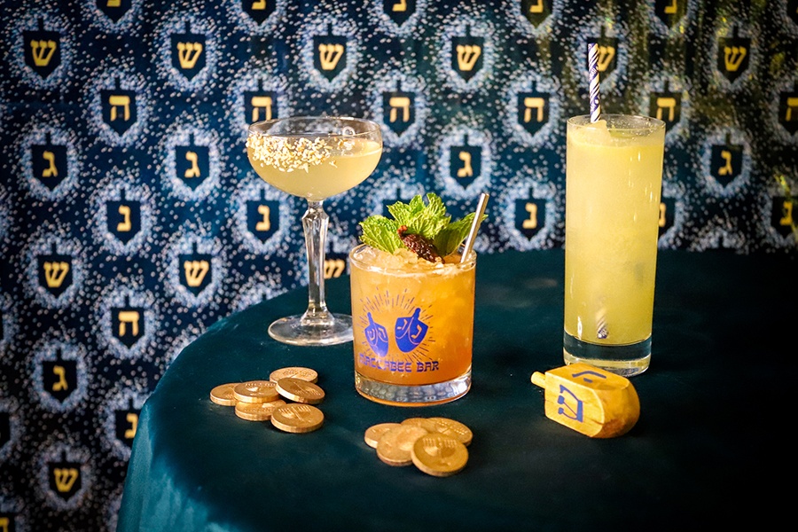 Three cocktails sit on a table surrounded by gelt and a dreidle, in front of Hanukkah-themed wallpaper.