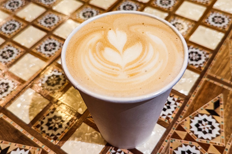 A latte in a paper cup is served on a table with an intricate checkers board built in.