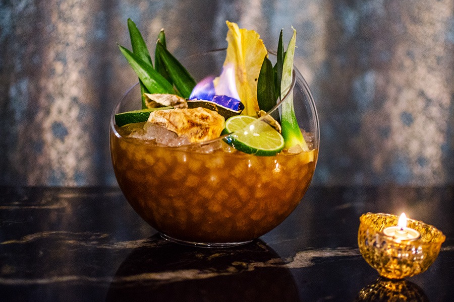 A drink is served in a large glass punchbowl with citrus, pineapple leaves, and something on fire.