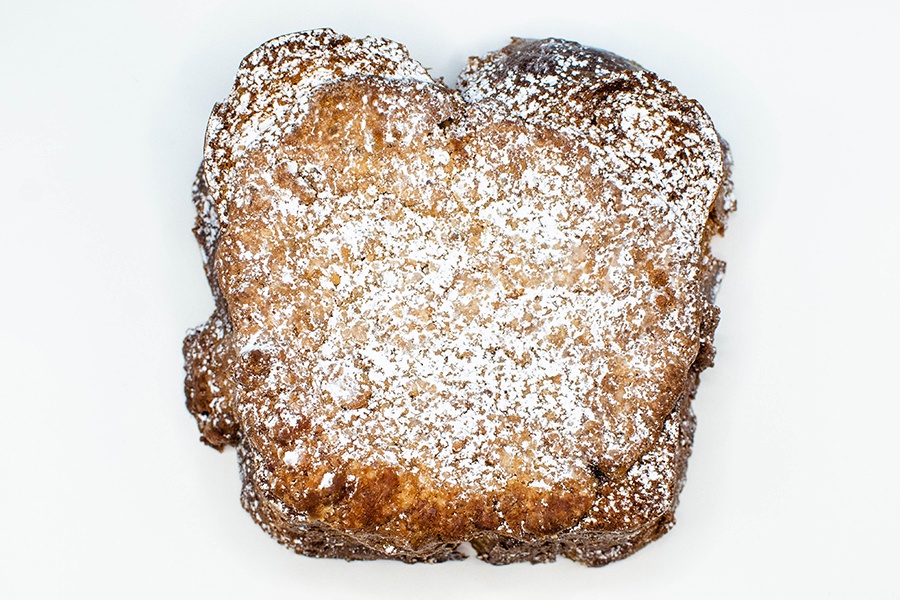 A piece of thick, baked French toast covered in powdered sugar sits on a white background.