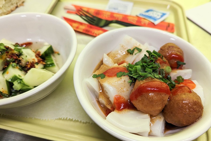 Two white bowls sit on a pale green tray, one featuring thick noodle rolls with fish balls and a swoosh of sriracha, the other with chopped cucumbers.
