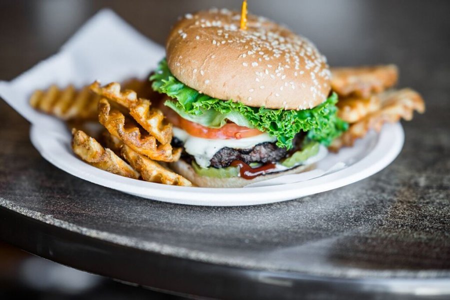 A burger with lettuce, tomato, cheese, and pickles is served on a white paper plate with waffle fries on the side.