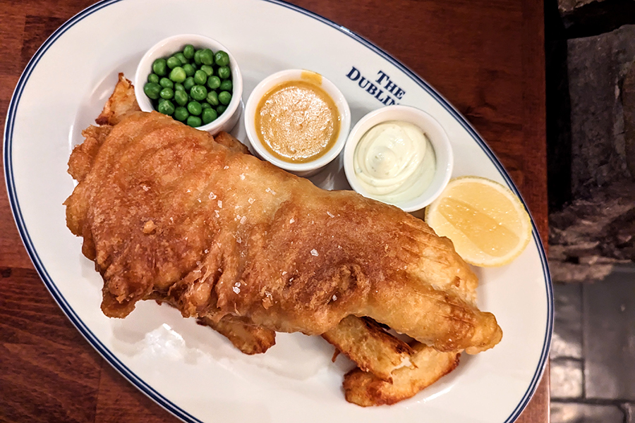 Overhead view of a large portion of fried haddock atop thick steak fries. Three small cups are on the side—one with peas, the other two with sauces.