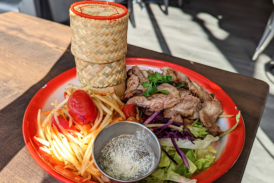 A white plate with a red rim holds generous portions of Thai papaya salad and pieces of grilled pork, along with a small woven basket holding sticky rice.