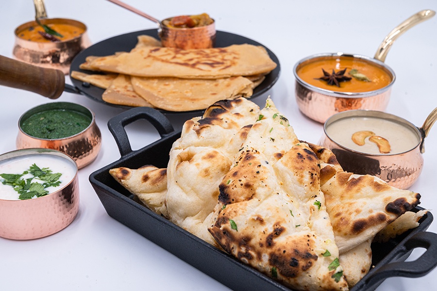 Two styles of Indian bread in cast-iron pans are isolated on a white background with five small copper pots of sauces.