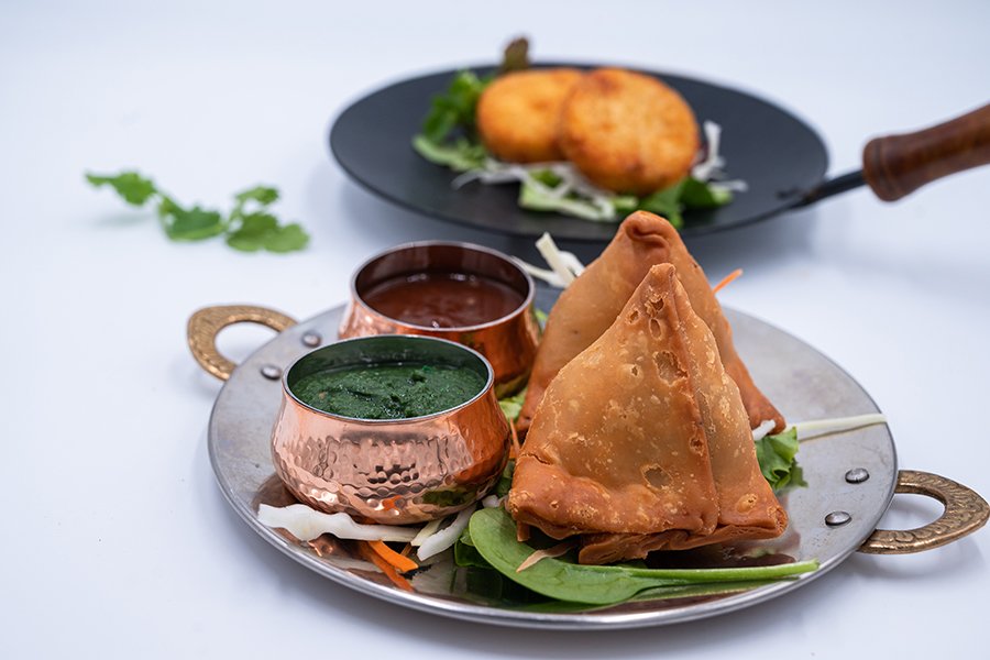 Two Indian samosas are displayed on a silver plate with small copper bowls of a green, herby sauce and a thick brown sauce. In the background, two fried potato patties are visible.