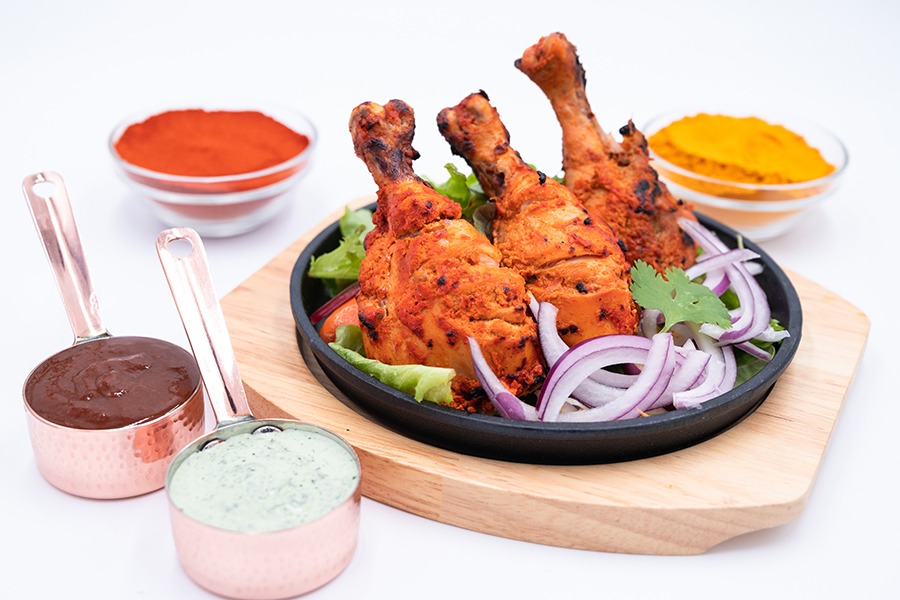 Three grilled bone-in chicken pieces are displayed on a black platter with sliced red onion, with bowls of sauces and spices visible around the plate.