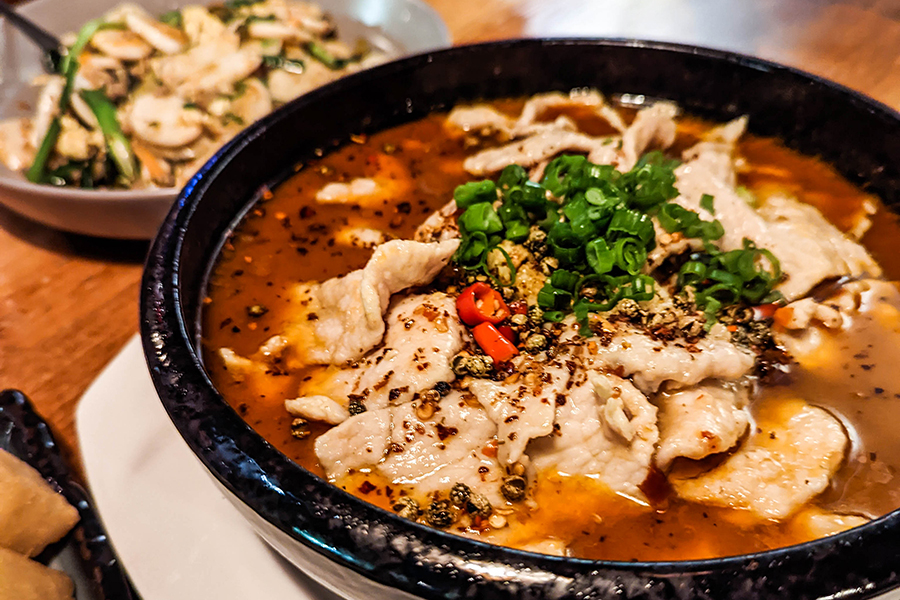 A big black bowl is full of sliced bowl and red broth garnished with plenty of chili pepper flakes and chopped scallions.