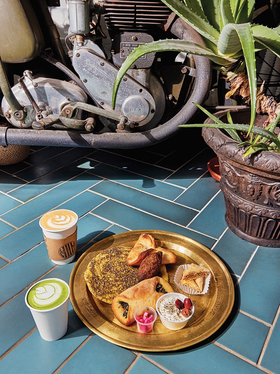 A golden tray sits on a teal tiled floor, holding Middle Eastern pastries, with a couple of lattes to the side.