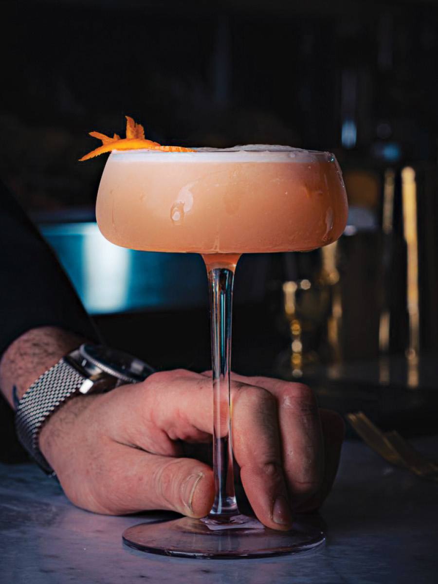 A hand holds a pale orange-pink cocktail in elegant glassware at a dark bar.