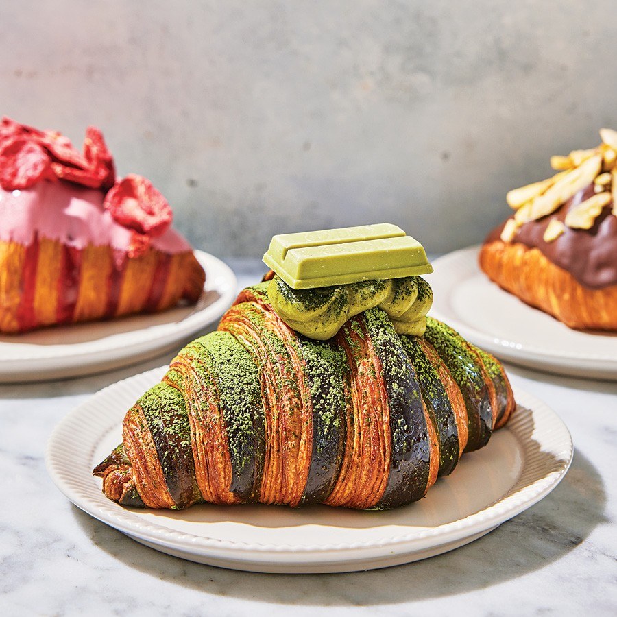 Three elaborate croissants with different toppings sit on white plates on a white marble surface.