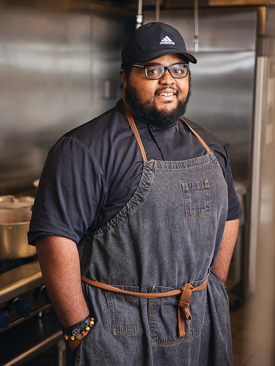 A bearded man in glasses and a baseball cap stands in a professional kitchen, wearing a denim apron and looking at the camera.