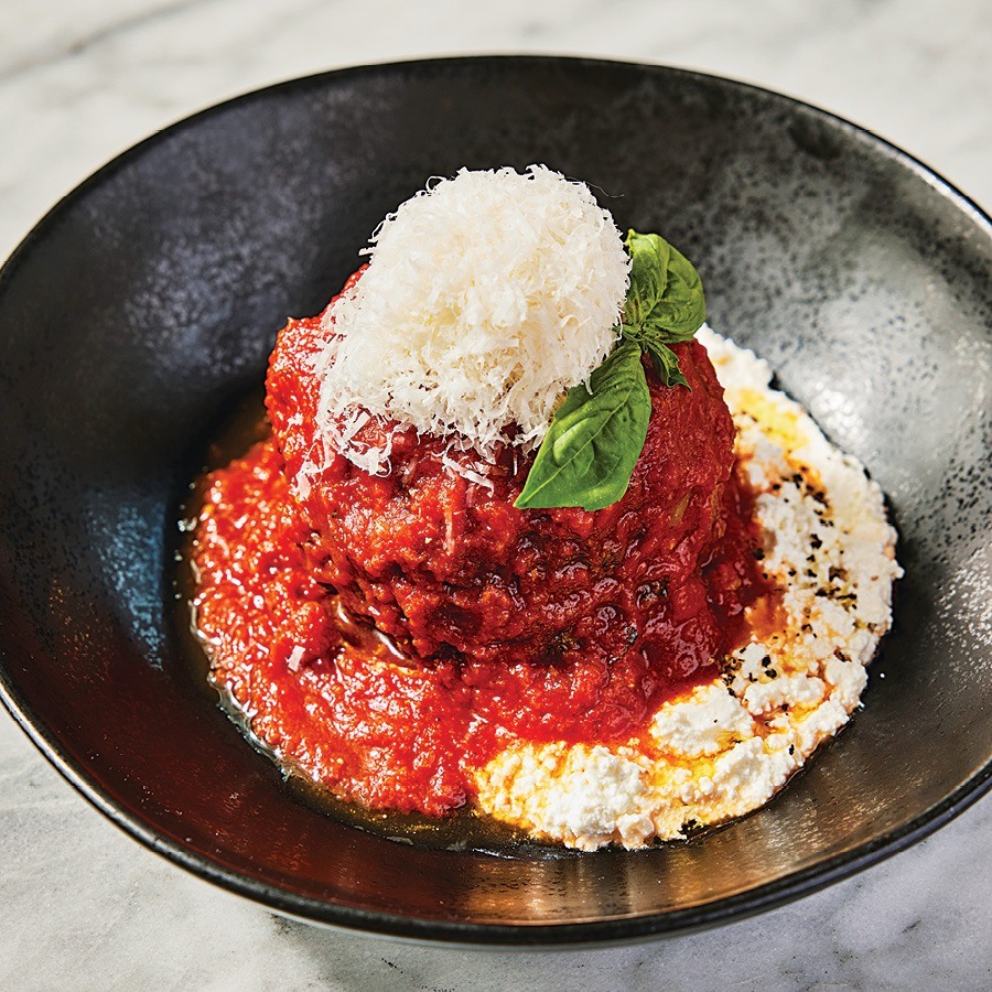 A giant, saucy meatball sits on a black plate, topped with a hefty amount of grated cheese and a basil leaf.