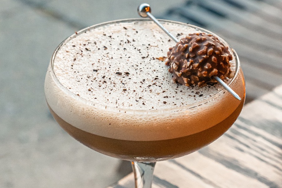 A light brown cocktail with a foamy top is garnished with a Ferrero Rocher bonbon.