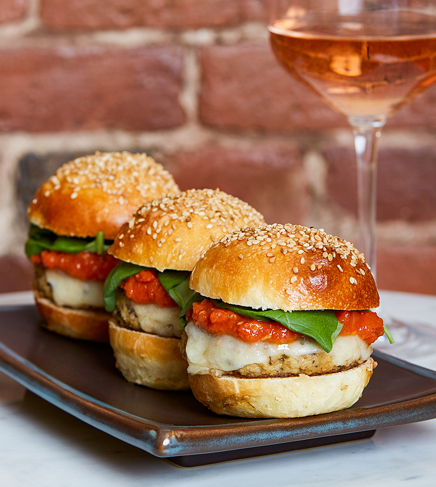 Three mini cheeseburgers topped with a tomato sauce and basil are displayed on a plate with a glass of rose to the side.