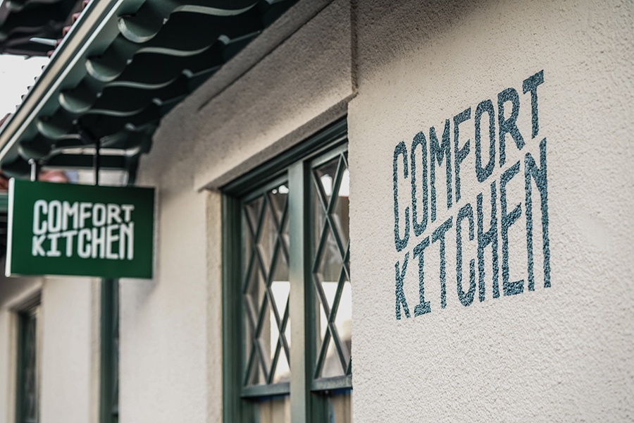 An exterior of a restaurant has the name, Comfort Kitchen, painted in green on the building, and a small green sign says the same.