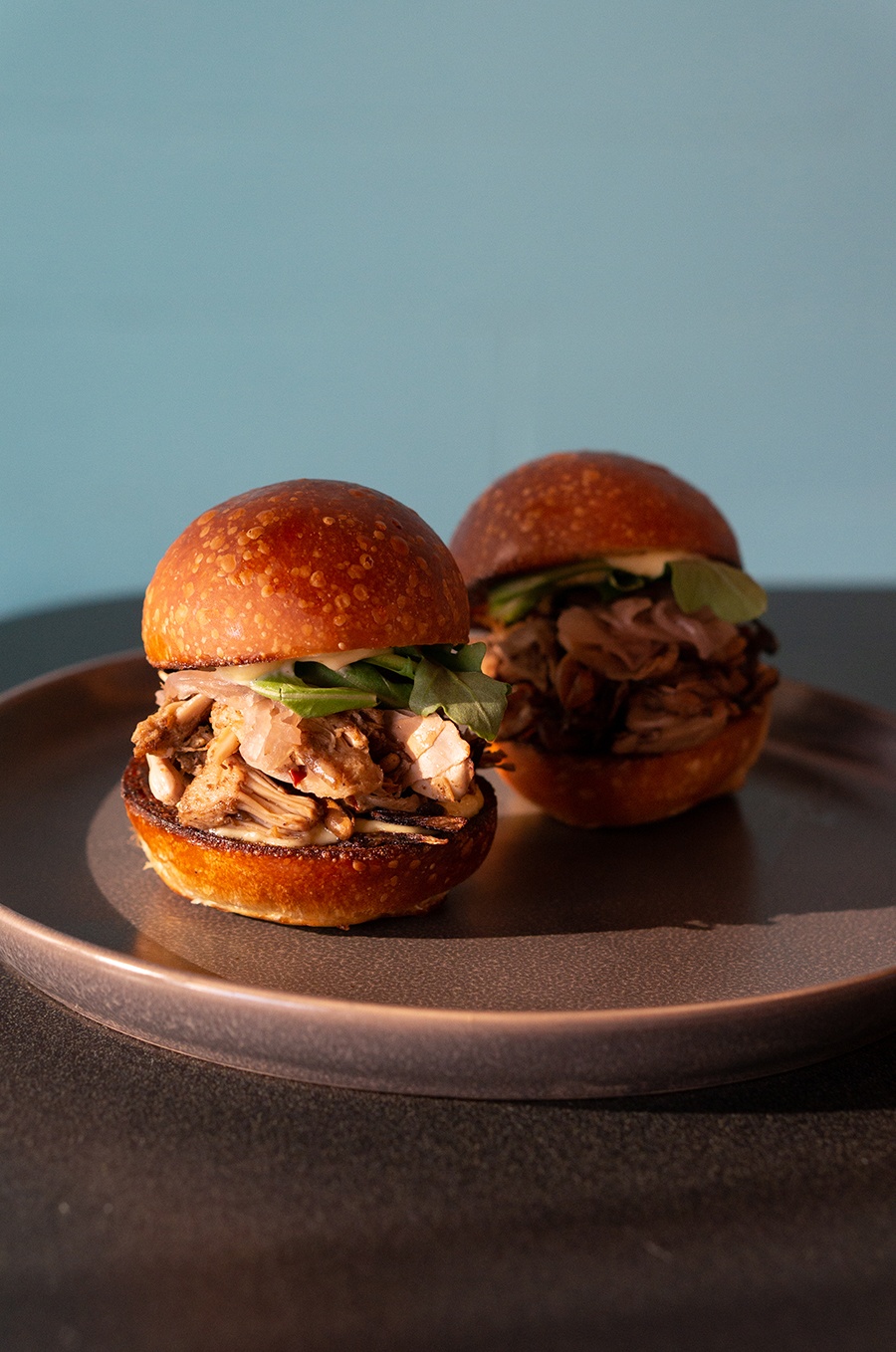 Two sliders sit on a plate, filled with a pulled meat substitute and arugula.
