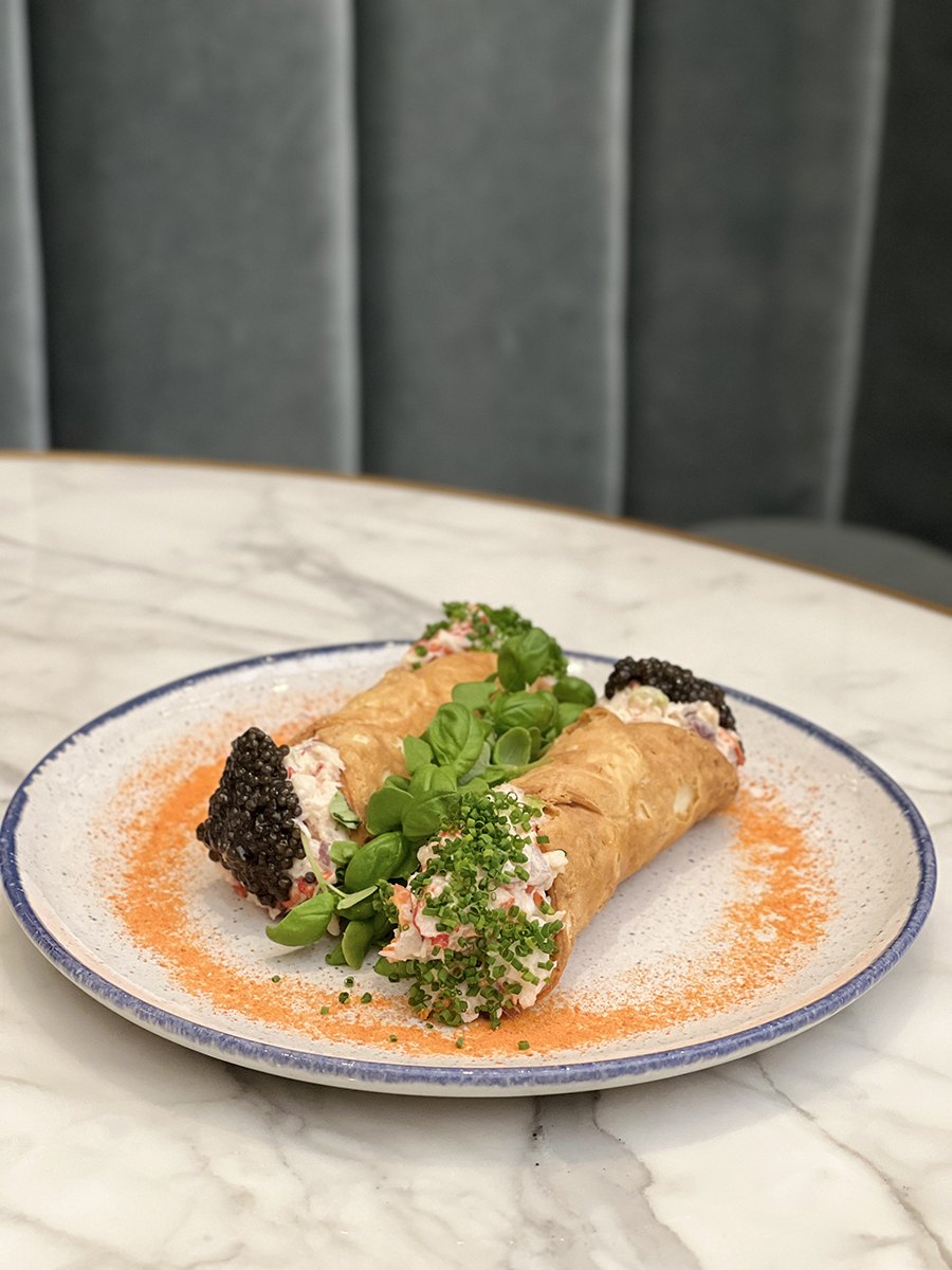 Two cannolis sit on a plate on a white marble table, each stuffed with lobster, chives, and caviar.
