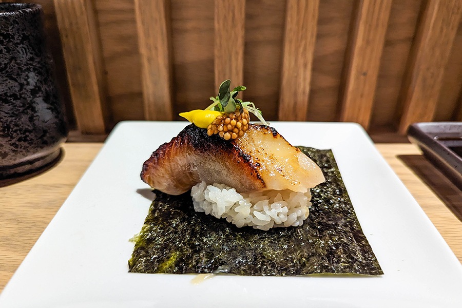 A piece of white fish with torched edges is topped with a dollop of whole mustard seeds, a bright yellow puree, and microgreens, and it sits on a ball of rice on seaweed.