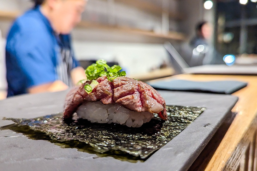 A rare piece of beef is draped over a ball of sushi rice atop a square of seaweed, sitting on a plate on a sushi bar.