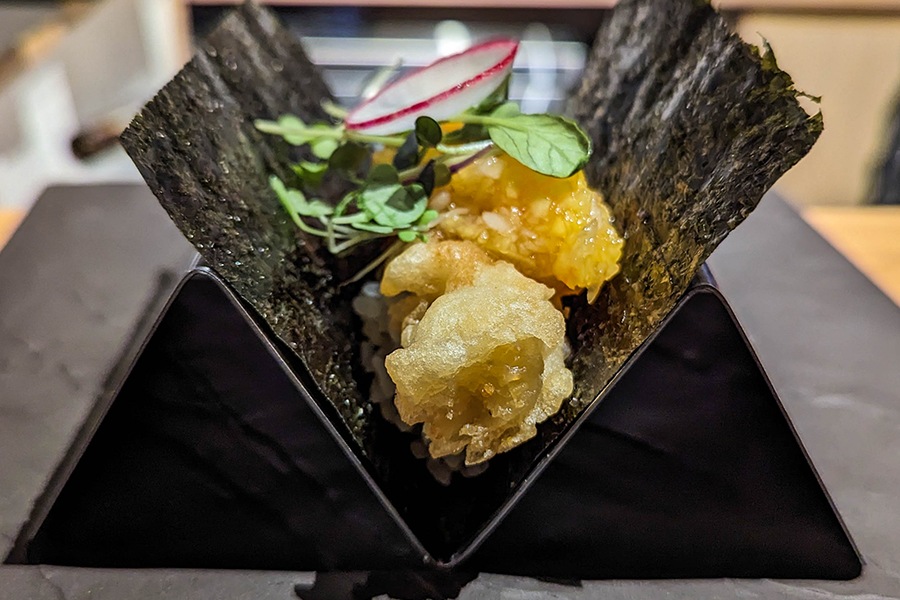 A sushi handroll is propped upright in a triangular taco stand, and it's filled with crispy shrimp, an orange salsa, microgreens, and a radish slice.
